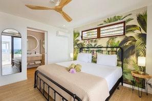 A hotel room with a bed, a ceiling fan, a bathroom with a mirror, and a tropical mural on the wall. - Spacious luxury villa with panoramic sea views (Sint Willibrordus)