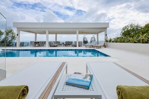 A poolside area with lounge chairs, a covered seating area, and a book on a table. - Spacious luxury villa with panoramic sea views (Sint Willibrordus)