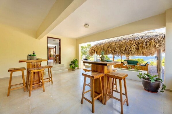Una zona de bar con barra de madera, taburetes, techo de paja y vista al océano.