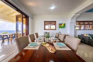 Un coin repas avec une table en bois dressée pour quatre personnes, entourée de chaises rembourrées, et offrant une vue sur l’océan grâce à de grandes fenêtres.