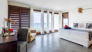 Un dormitorio moderno con una cama grande, un escritorio con una computadora portátil, una silla y vista al mar a través de ventanas amplias.