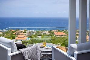A balcony with a view of the ocean, a round table with drinks, and a sofa. - Design villa with sea views and infinity pool (Sint Willibrordus)