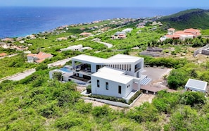 A modern white building with a pool, surrounded by lush greenery and a coastal view. - Design villa with sea views and infinity pool (Sint Willibrordus)