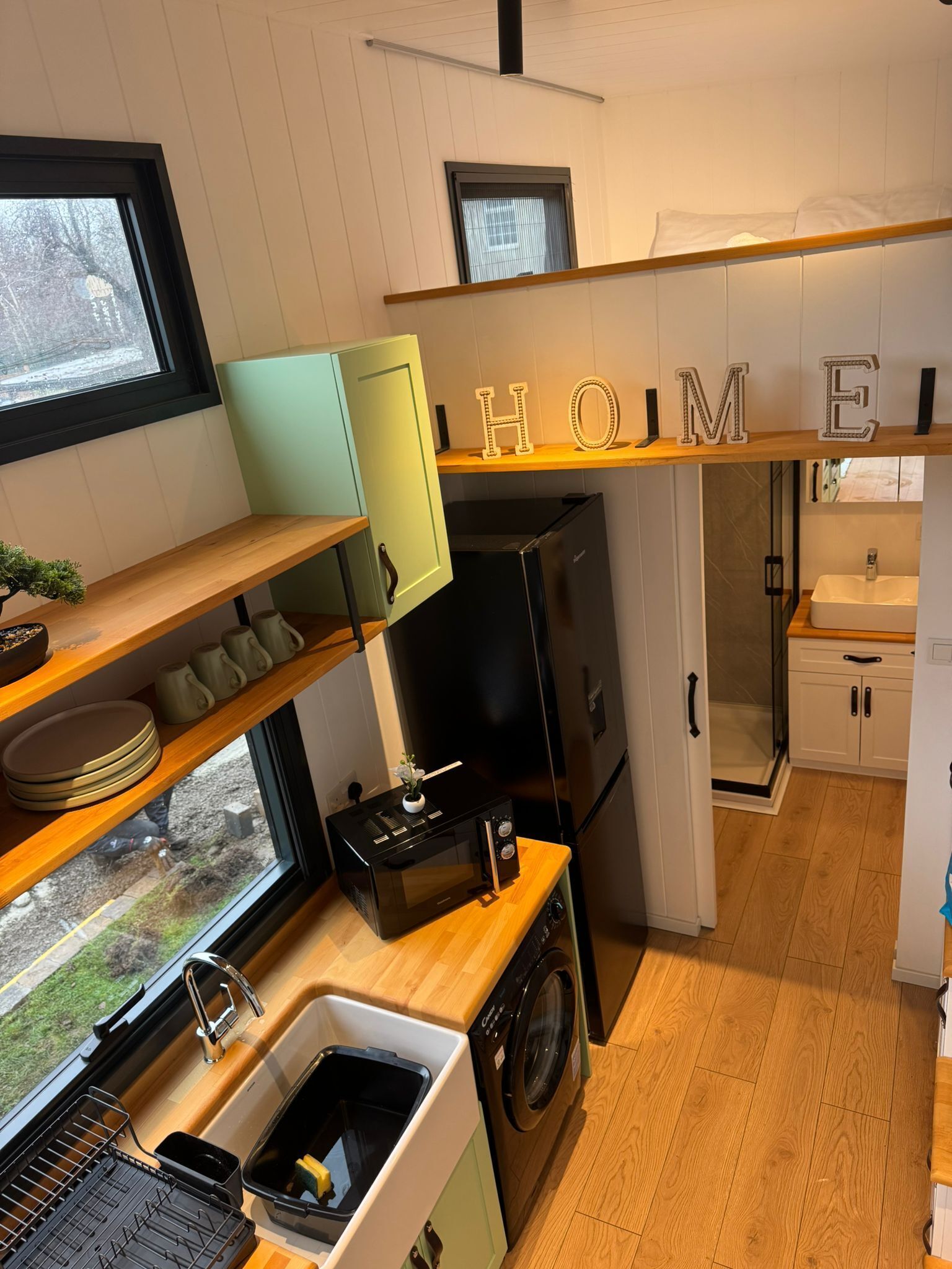 Cocina compacta con microondas, horno y lavaplatos. Hay estantes de madera y una ventana con vistas al exterior.
