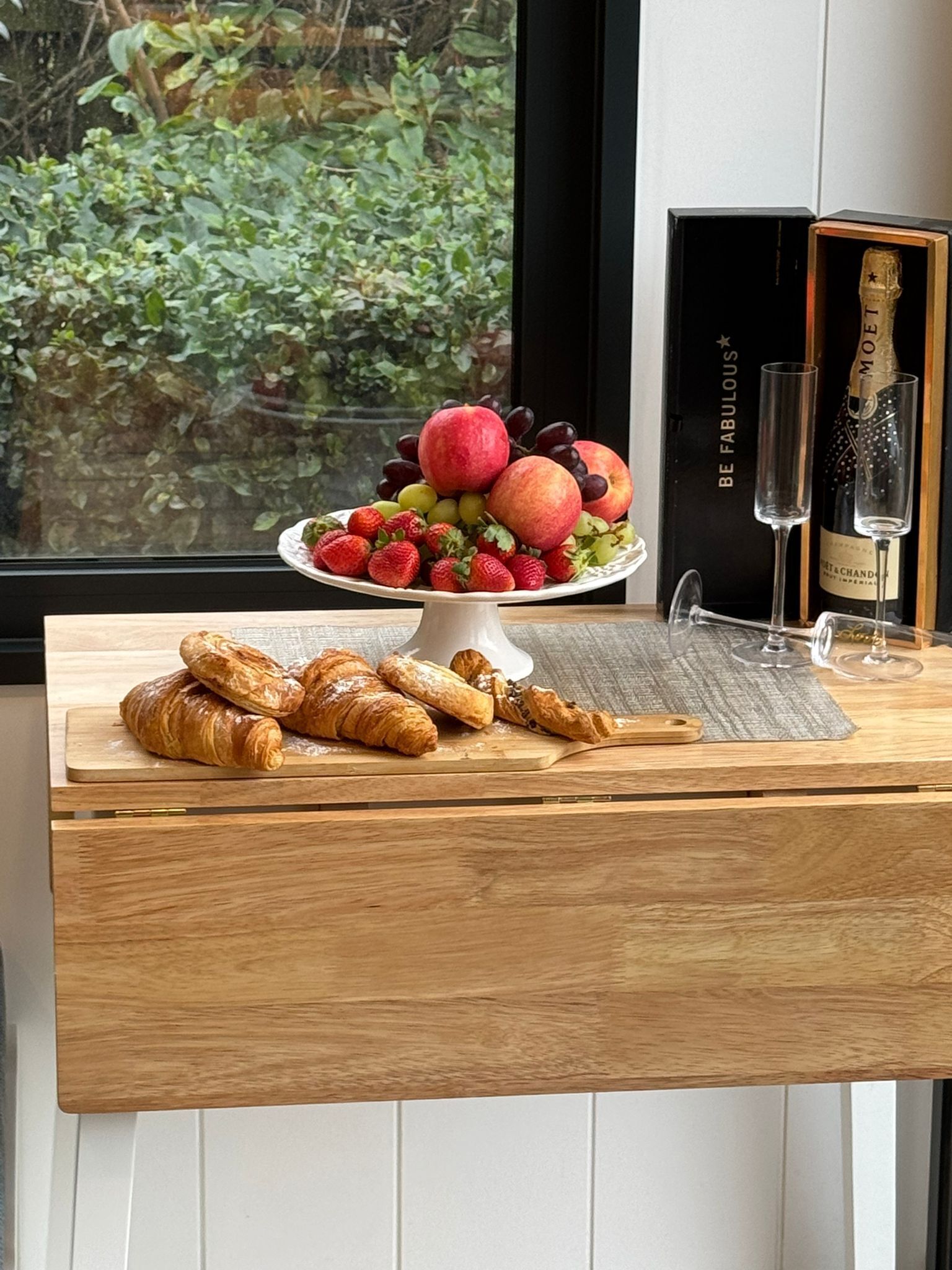A wooden table with a fruit platter, two glasses, and a bottle of champagne.