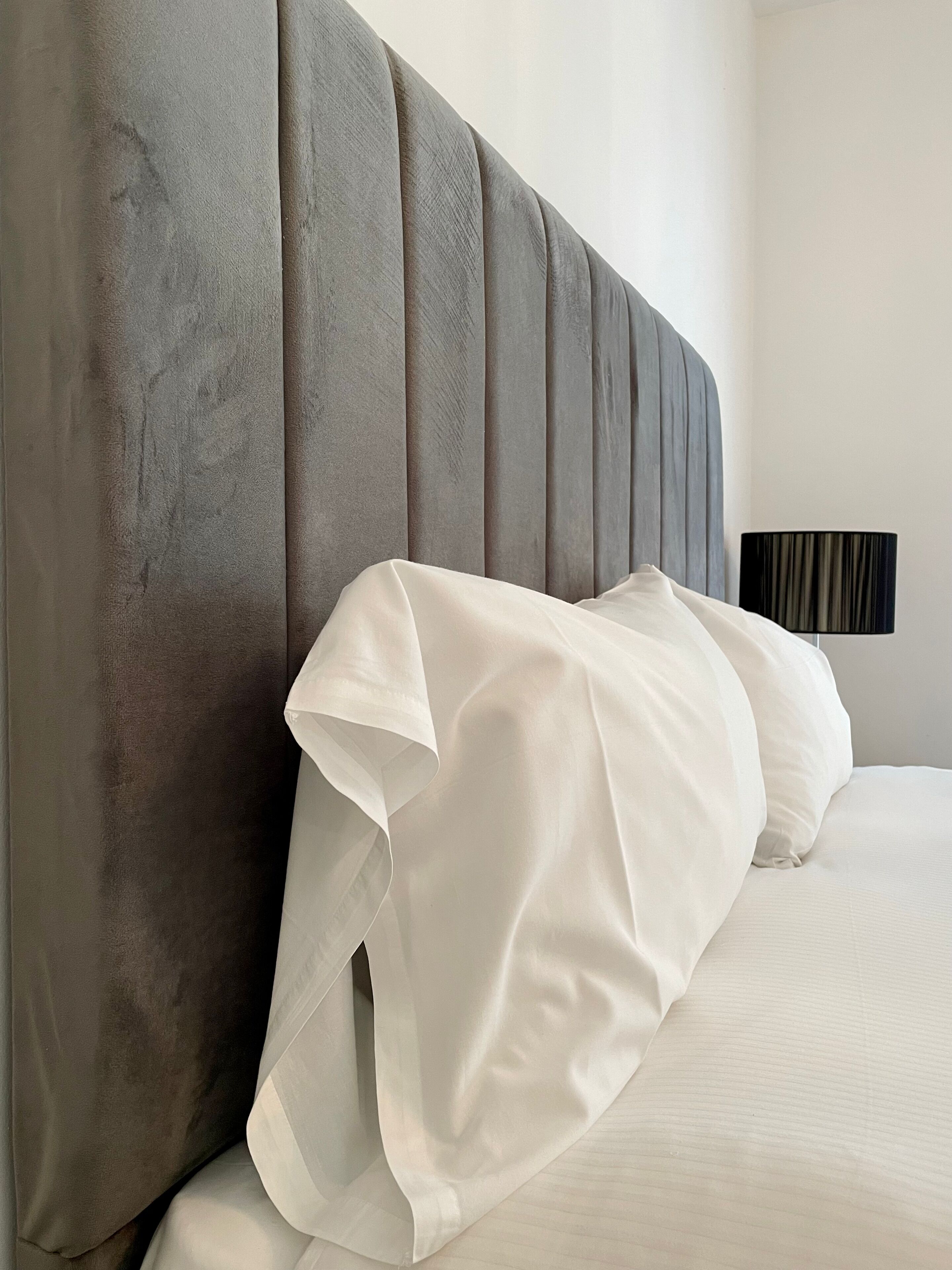 Un lit avec du linge de lit blanc et une tête de lit rembourrée de couleur grise.