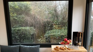 A cozy room with a large window, a wooden bench, a small table with food and drinks, and a view of greenery outside. - Old Mill Tiny Homes (Yarm)