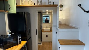 A compact kitchen with a microwave, toaster, and washing machine. A wooden staircase leads to a bathroom with a sink and mirror. - Old Mill Tiny Homes (Yarm)