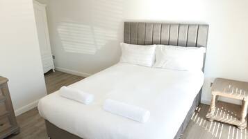 Una cama bien tendida con sábanas blancas y dos toallas enrolladas, una mesita de noche de madera y un taburete pequeño de madera.