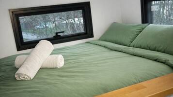 A single bed with green bedding and a wooden headboard, two windows with black frames, and a view of trees outside.