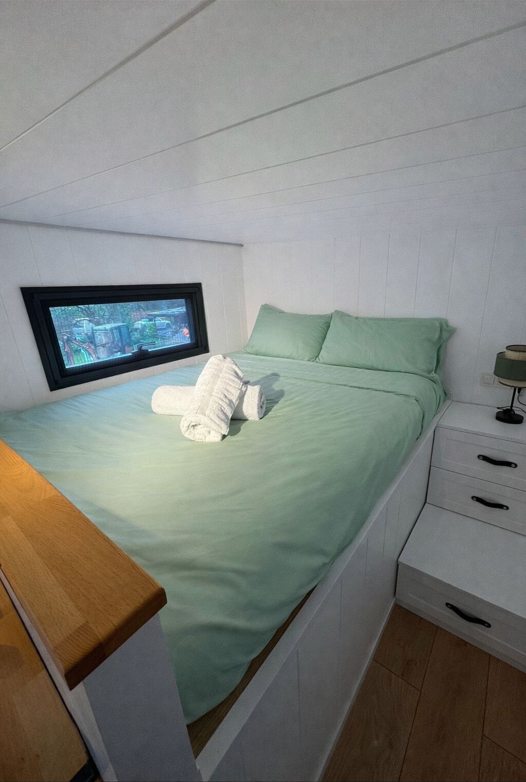 A single bed with a green bedspread and pillows, a wooden nightstand with a lamp, and a television mounted on the wall.
