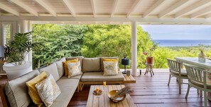 A covered outdoor seating area with a sofa, dining table, and chairs, overlooking a lush landscape and the ocean. - Spacious villa with pool and ocean views (Sint Willibrordus)