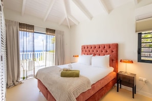 A bedroom with a large bed, a nightstand, a lamp, and a view of the sea through the window. - Designer sea-view villa with private pool (Sint Willibrordus)