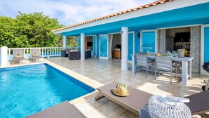Zona de piscina con mesa y sillas, una silla de descanso y una mesita con un sombrero.