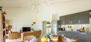 A modern kitchen with dark cabinets, a dining area with a wooden table set for two, and a living area with a sofa and a television. - Modern villa with private pool and garden views (Sint Willibrordus)