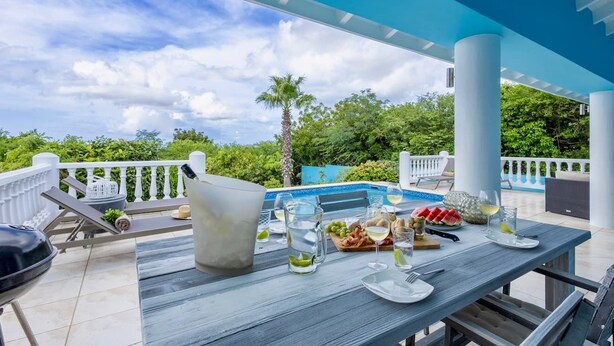 Un área de comedor exterior con vista a la piscina, una parrilla y una mesa lista para una comida.