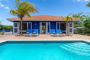 Uma área de piscina com piscina, espreguiçadeiras azuis e um edifício com telhado em azulejos.