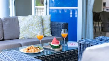 A breakfast setup with croissants, watermelon slices, and orange juice on a table, with a sofa and wicker furniture in the background.
