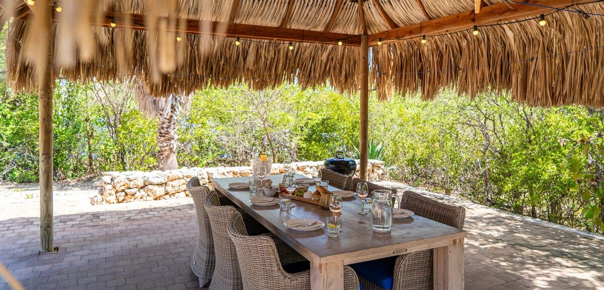 An outdoor dining area with a thatched roof, wicker furniture, and a stone wall in the background.