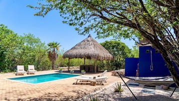 A swimming pool with lounge chairs, a thatched-roof hut, and a blue building.