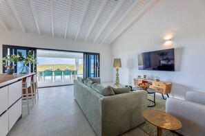 A modern living room with a sofa, a coffee table, and a TV mounted on the wall. There is a dining area with chairs and a table, and a kitchen area with cabinets and a countertop. - Modern villa with infinity pool and sea views (Sint Willibrordus)