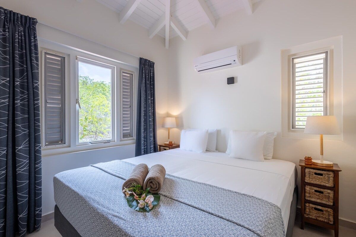 A hotel room with a bed, two bedside tables, a window with shutters, and an air conditioning unit.