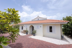A single-story building with a tiled roof, a brick pathway leading to the entrance, and a small porch area with a bench and potted plants.