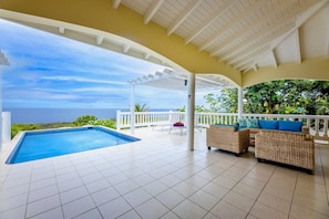 A covered outdoor area with a swimming pool, wicker furniture, and a view of the ocean.