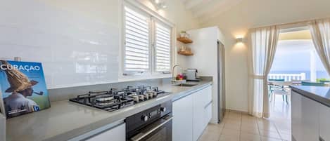 A modern kitchen with a gas stove, oven, and white cabinetry. There is a window with blinds, a countertop with a sink, and a view of the outdoors.