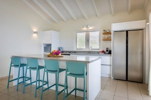 A modern kitchen with a central island, bar stools, a stainless steel refrigerator, and a window with blinds.