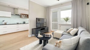A modern living room with a grey sofa, a black coffee table, and a TV on a black stand. There is a kitchen in the background with white cabinets and a wooden countertop. - Comfortable Apartment in Poznan | Garage | Balcony (Poznań)