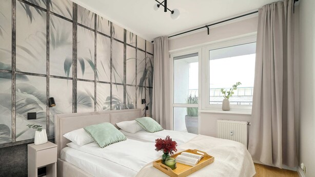A modern bedroom with a large bed, a bedside table, a window with curtains, and a view of the outdoors. - Comfortable Apartment in Poznan | Garage | Balcony (Poznań)