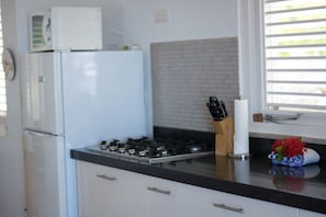 Una cocina con un refrigerador blanco, una estufa y un microondas encima. Hay armarios con manijas, una ventana con persianas y un juego de cuchillos en la encimera.