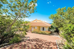 Una casa amarilla con techo de tejas, rodeada de vegetación y un sendero pavimentado.