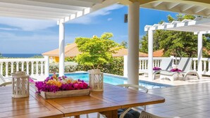 A covered outdoor area with a wooden table, white railings, and a view of a swimming pool and ocean. - Elegant sea-view villa with private pool - Coral Estate (Sint Willibrordus)