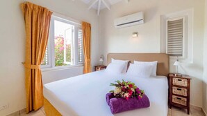 A hotel room with a large bed, a window with curtains, an air conditioning unit, a bedside table with a lamp, and a small wooden nightstand. - Elegant sea-view villa with private pool - Coral Estate (Sint Willibrordus)