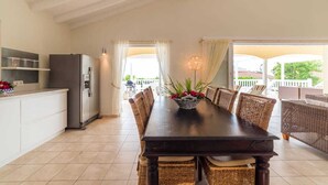 A dining area with a dark wooden table, wicker chairs, and a basket of flowers. A modern kitchen with a refrigerator and cabinets is visible to the left. - Elegant sea-view villa with private pool - Coral Estate (Sint Willibrordus)