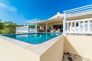 Una piscina con una pérgola blanca y sillas de descanso, rodeada por un edificio de color beige con techo de tejas rojas.