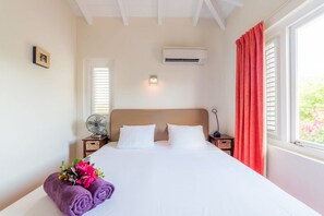 Habitació d'hotel amb un llit, aire condicionat fixat a la paret, ventilador al costat del llit i una finestra amb cortines vermelles.
