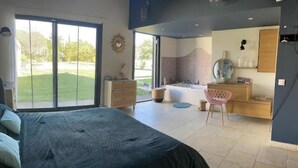 A modern bedroom with a large bed, a bathroom with a bathtub, and a view of a green lawn. - Provencal Escape: Villa with Pool and Pool House in the Heart of the Luberon (La Tour-d'Aigues)