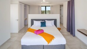 A modern bedroom with a large bed, a grey headboard, and a view of the outdoors through a window. - Luxury sea view villa with private pool (Sint Willibrordus)