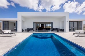 A modern outdoor swimming pool area with a clear sky, two lounge chairs, and a seating arrangement. - Luxury sea view villa with private pool (Sint Willibrordus)