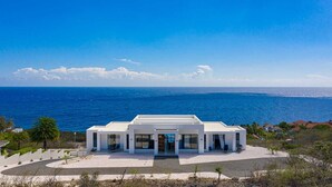 Un edificio moderno de color blanco con techo plano, amplios ventanales y una entrada central. Se encuentra sobre una zona de arena blanca con vistas al océano y un cielo azul despejado.