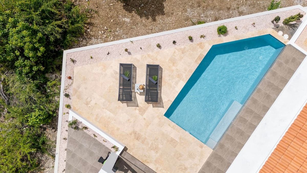 An aerial view of a swimming pool area with two sun loungers and a small table, surrounded by a pebble beach and greenery.