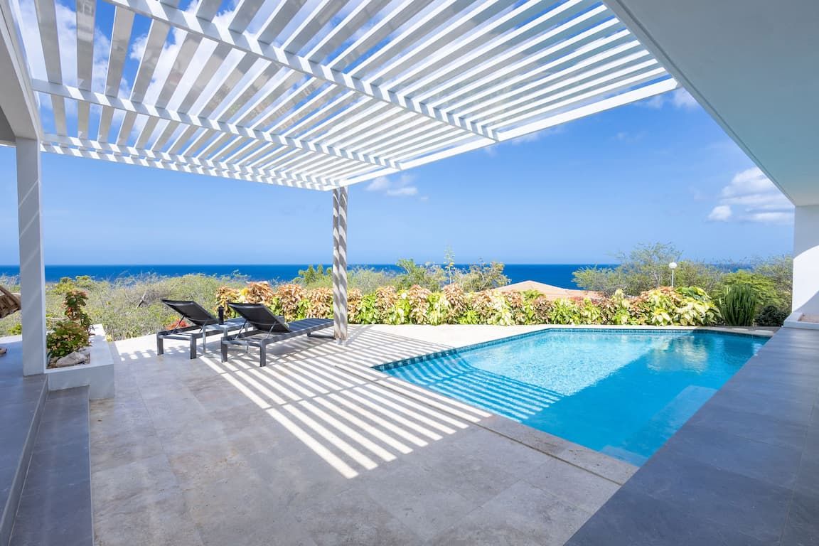Ein moderner Poolbereich im Freien mit Blick auf den Ozean, eine Pergola spendet Schatten, und einige Liegestühle.