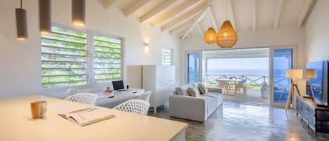 A spacious living area with a large window overlooking a scenic view, a white sofa, a dining table, and a kitchen area with a laptop and a coffee cup.