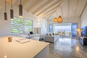 A spacious living area with a large window overlooking a scenic view, a white sofa, a dining table, and a kitchen area with a laptop and a coffee cup. - Romantic garden bungalow with pool and sea view (Sint Willibrordus)