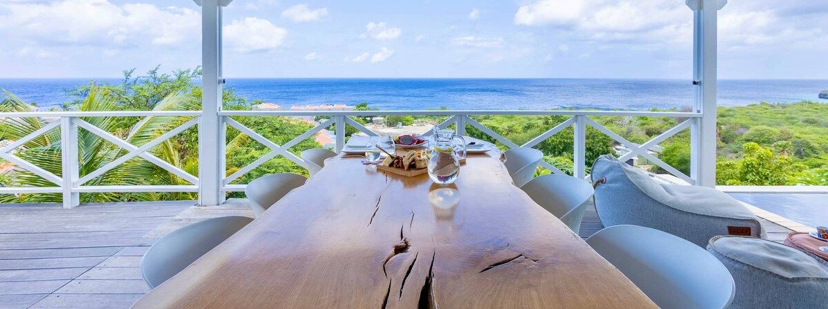 Een houten tafel met stoelen, een witte leuning en uitzicht op de oceaan.