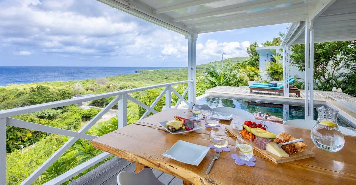 A dining table set with food and drinks, overlooking a pool and ocean view.