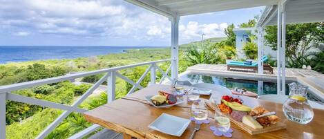 A dining table set with food and drinks, overlooking a pool and ocean view.